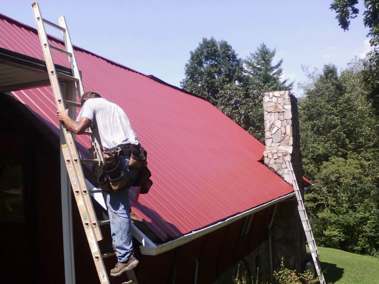 Roofer on a ladder beside a red metal roof and stone chimney — Soffit & Fascia Repair by Roofing Brothers in Buchanan